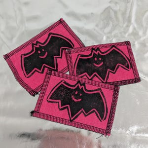May include: Three pink fabric patches featuring a black bat design with a smiling face. The rectangular patches have a black stitched border. They are arranged on a shiny silver surface.
