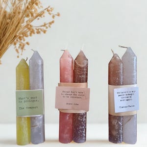 May include: Three sets of paired pillar candles in shades of yellow, silver, pink, brown, and gray. Each set is wrapped with a paper band featuring quotes from The Tempest, Steve Jobs, and Thomas Paine. The candles have a textured, mottled appearance.