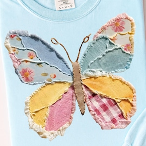 May include: Light blue t-shirt featuring a colorful butterfly applique. The butterfly wings are patchwork, with sections of pink, yellow, blue, and floral patterns. The shirt has a crew neck and short sleeves.