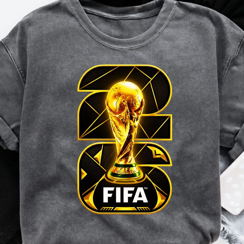 以下が含まれることがあります： 金色のFIFAワールドカップトロフィーのデザインが施された濃いグレーのTシャツ。デザインにはFIFAのロゴと、黒と金の幾何学模様で描かれた数字の23が含まれています。トロフィーはシャツの中央に配置されています。