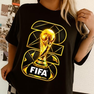 以下が含まれることがあります： 金色のFIFAワールドカップトロフィーのデザインが施された黒いTシャツ。デザインにはFIFAのロゴと2022の数字が幾何学的なスタイルで含まれています。このシャツは半袖のクルーネックです。