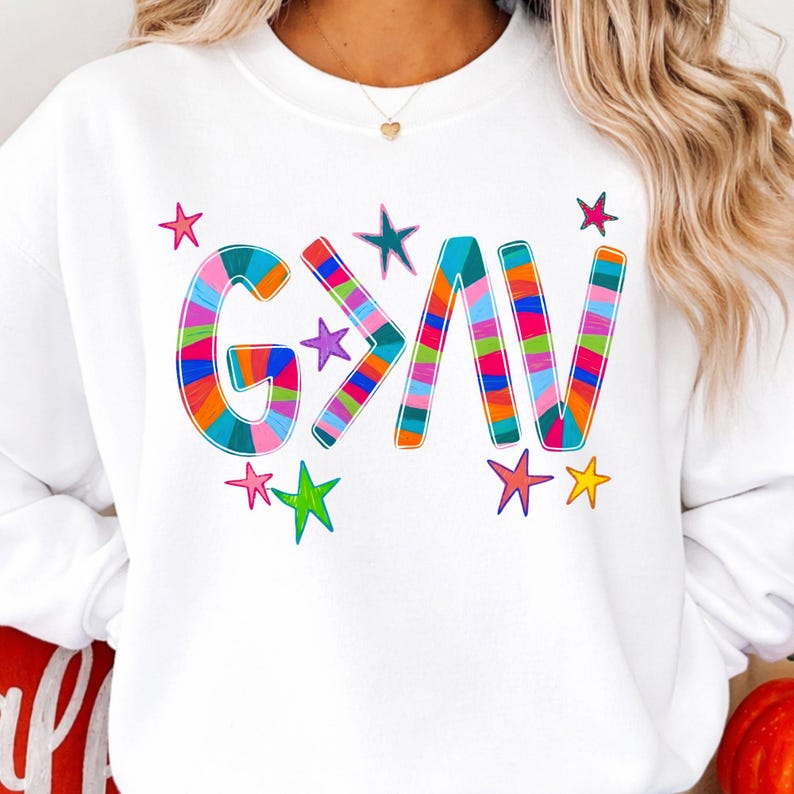 以下が含まれることがあります： カラフルなストライプ文字で「G>AV」と書かれた白いスウェットシャツ。テキストの周りには様々な色の星がデザインされています。このスウェットシャツはカジュアルな普段着です。