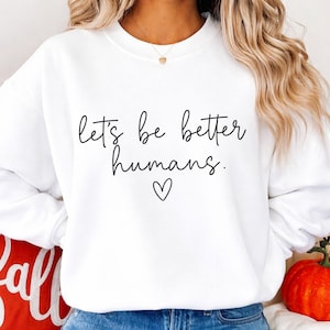 May include: White sweatshirt with the phrase "let's be better humans" in black script, with a small heart below the text. The sweatshirt is a simple, casual style.