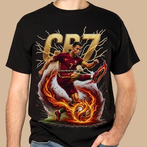 May include: Black t-shirt featuring a dynamic graphic of a soccer player in a red and green uniform kicking a flaming soccer ball. The design includes the text "CR7" in gold, with lightning bolt accents.
