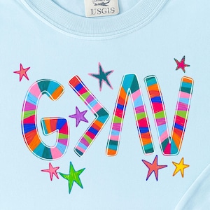 以下が含まれることがあります： 水色のスウェットシャツ。カラフルな虹色のストライプ文字で「G>AV」と書かれています。ピンク、紫、緑、黄色の星の形がデザインされています。スウェットシャツはクルーネックで、「USGIS」と書かれたタグが付いています。