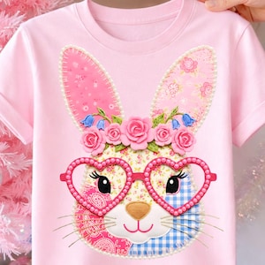 Pode incluir: T-shirt rosa de manga curta com um design de rosto de coelho. O coelho tem óculos em forma de coração, uma coroa de flores e um design de patchwork com padrões rosa, azul e florais. As orelhas do coelho são rosa com detalhes florais.