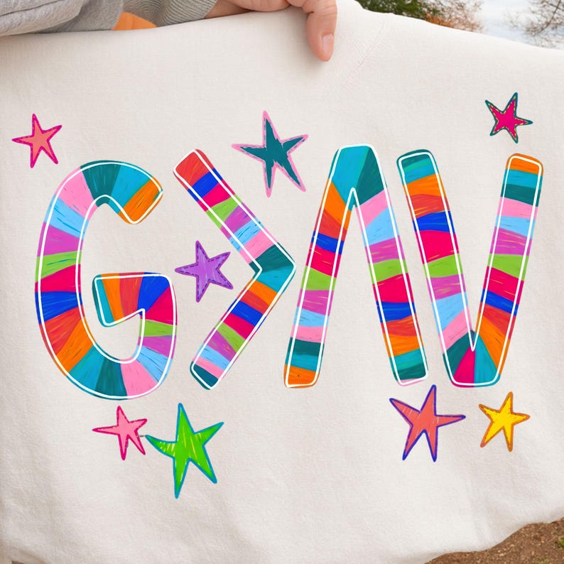 以下が含まれることがあります： カラフルなストライプの文字で「G>N」と書かれた白いスウェットシャツ。文字は白で縁取られ、様々な色の星の形に囲まれています。手描きのデザインです。