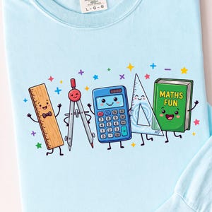 Peut inclure: Un t-shirt à manches longues bleu clair avec un motif de cinq fournitures scolaires souriantes : une règle en bois avec un nœud papillon, un compas, une calculatrice bleue, une équerre transparente et un livre vert avec le texte "MATHS FUN".