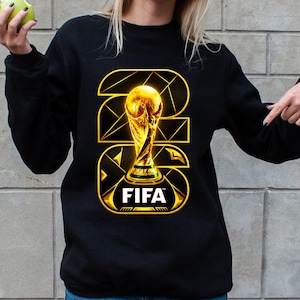 以下が含まれることがあります： 金色のFIFAワールドカップトロフィーのデザインが施された黒いスウェットシャツ。デザインには、金色の輪郭で「22」と「26」の数字と、FIFAのロゴが含まれています。スウェットシャツは無地の黒色です。