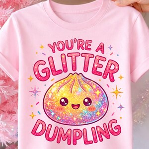 Può includere: T-shirt rosa con la scritta "You're a Glitter Dumpling". Il design presenta un raviolo glitterato con un volto sorridente, circondato da stelle e brillantini. Il testo e il raviolo sono rosa e glitterati.