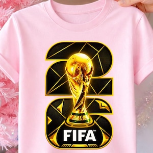 以下が含まれることがあります： 薄いピンク色のTシャツ。金色のFIFAワールドカップトロフィーのデザインが施されています。グラフィックには、黒と金の幾何学模様を背景に、金と白で「FIFA」の文字が含まれています。Tシャツは半袖です。