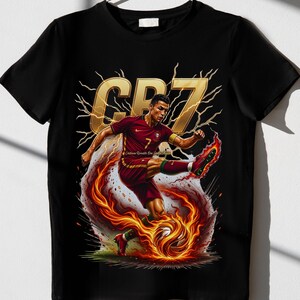 May include: Black t-shirt featuring a dynamic illustration of a soccer player in a maroon uniform kicking a flaming soccer ball. The design includes the gold letters "CR7" and lightning bolt accents.