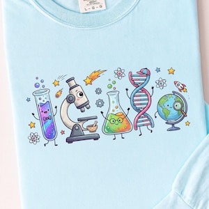 Può includere: Una maglietta a maniche lunghe azzurra con una stampa grafica colorata di attrezzature scientifiche animate, tra cui una provetta, un microscopio, un becher, un filamento di DNA e un mappamondo, circondati da piccole stelle, atomi e un razzo.