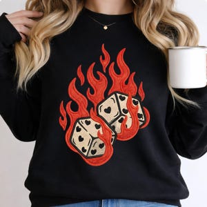 May include: Black sweatshirt featuring an embroidered design of two dice engulfed in flames. The dice are tan with black heart-shaped pips. The flames are bright red, creating a bold contrast against the black fabric. A casual, comfortable garment.