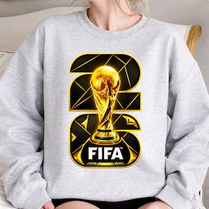 以下が含まれることがあります： 金色のFIFAワールドカップトロフィーのデザインが施された、明るい灰色のスウェットシャツ。デザインにはFIFAのロゴと2026年が黒と金の幾何学模様で描かれています。トロフィーは中央に配置され、目立っています。