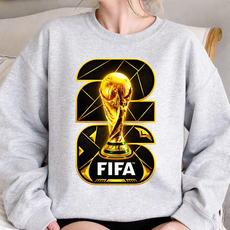 以下が含まれることがあります： 金色のFIFAワールドカップトロフィーのデザインが施された、明るい灰色のスウェットシャツ。デザインにはFIFAのロゴと2026年が黒と金の幾何学模様で描かれています。トロフィーは中央に配置され、目立っています。