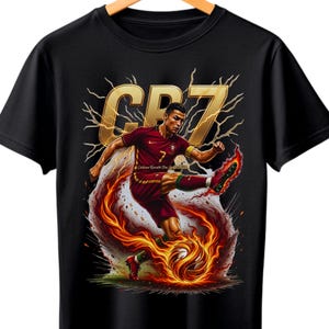May include: Black t-shirt featuring a graphic of a soccer player in a maroon uniform kicking a soccer ball engulfed in flames. The design includes the letters "CR7" in gold with lightning bolts.