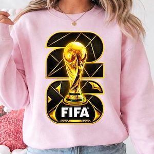 以下が含まれることがあります： 金色のFIFAワールドカップトロフィーのデザインが施されたピンクのスウェットシャツ。デザインには、黒と金の幾何学模様で23の数字と、下部にFIFAのロゴが含まれています。スウェットシャツは明るいピンク色です。