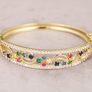 May include: A gold-coloured bangle bracelet with a decorative design. The bracelet features a row of small, clear stones and colourful gemstones in blue, green, and pink. The bracelet has an intricate, openwork pattern.