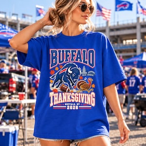 Könnte beinhalten: Ein königsblaues Kurzarm-T-Shirt mit einem Grafikdruck, der einen anstürmenden Büffel, einen Football, einen Truthahn und Herbstblätter zeigt. Der Text lautet BUFFALO THANKSGIVING, BILLS COUNTRY sowie BUFFALO, NY 2026 in weißer und roter Schrift.