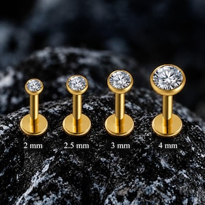 Könnte beinhalten: Vier goldfarbene Labret-Stecker mit klaren, runden Steinen in Zargenfassung, von links nach rechts nach Größe angeordnet. Die Stecker sind mit den Größen 2 mm, 2,5 mm, 3 mm und 4 mm beschriftet, die den Durchmesser der Steinfassung angeben.