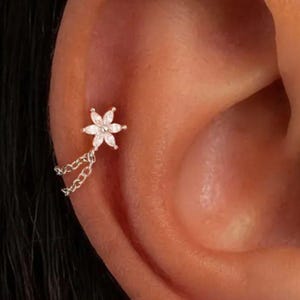 Könnte beinhalten: Ein silberfarbenes Ohrpiercing mit einem sechsblättrigen Blumenstecker, der mit klaren, marquise-geschliffenen Steinen besetzt ist. Zwei zarte silberne Ketten verlaufen von der Basis der Blume zu einem zweiten Befestigungspunkt am Ohr.
