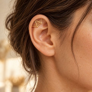 Può includere: Un ear cuff dorato a forma di ragnatela, caratterizzato da un delicato design traforato con sottili linee metalliche che si irradiano da un punto centrale. Il gioiello è posizionato lungo la cartilagine superiore dell'orecchio, seguendone la curva.