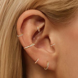 Pode incluir: Uma coleção de cinco argolas douradas usadas em vários piercings de orelha. Cada argola apresenta uma banda fina cravejada com pequenas pedras claras e brilhantes. Os brincos estão posicionados no lóbulo, hélix, rook e tragus.
