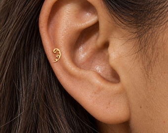 May include: A small gold-colored earring shaped like a delicate, curved scroll or filigree vine. The piece is positioned along the outer edge of the ear cartilage, featuring a polished metallic finish and a slender, tapering design with a spiral end.