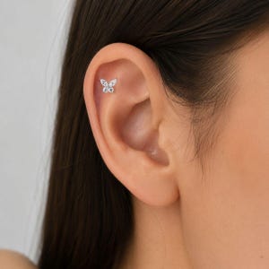 Può includere: Un piccolo orecchino a lobo a forma di farfalla color argento con quattro pietre trasparenti e scintillanti che formano le ali. Il gioiello è posizionato nella parte superiore della cartilagine dell'orecchio, con un design delicato.