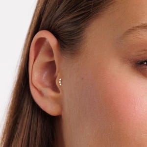 May include: A gold-toned cartilage earring featuring three small, clear, round-cut stones arranged in a vertical line. The earring is positioned in the tragus area of the ear, showcasing a delicate, minimalist design with a polished metal finish.