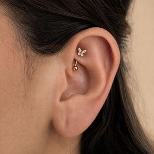 May include: A gold rook piercing jewellery piece featuring a butterfly-shaped charm adorned with small clear stones. The barbell has a smooth gold ball at the bottom end and the butterfly design at the top, positioned within the upper ear cartilage.
