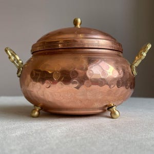 May include: A round copper bowl with a matching lid, featuring a hammered texture across the surface. The vessel includes two ornate brass side handles and stands on three small, spherical brass feet, providing a stable base for the container.
