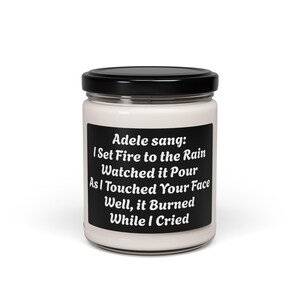 May include: A white candle in a glass jar with a black lid. The candle has a black label with white text that reads "Adele sang: I Set Fire to the Rain Watched it Pour As I Touched Your Face Well, it Burned While I Cried."