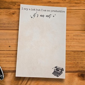 Puede incluir: Bloc de notas con el texto "I cry a lot but I am so productive, it's an art" y una ilustración de máquina de escribir. El bloc de notas está sobre un escritorio de madera con un bolígrafo, una cámara y una taza de café.