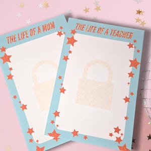 May include: Two rectangular notepads with light blue borders and red star accents. The text "THE LIFE OF A MOM" and "THE LIFE OF A TEACHER" are printed on the top. Each notepad has a light beige lock graphic in the centre. The background is pink with star confetti.
