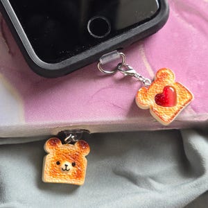 May include: Two phone charms, one shaped like a slice of toast with a red heart and the other shaped like a bear-faced toast. Both charms are attached to a phone case via a silver-colored metal loop. The phone case is black.