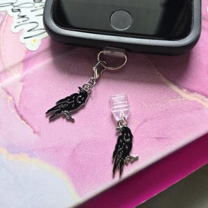 May include: Black raven phone charm with a silver-colored clasp and a clear phone plug. The raven charm has a crescent moon design. The phone is in a black case, resting on a pink and white marbled surface.