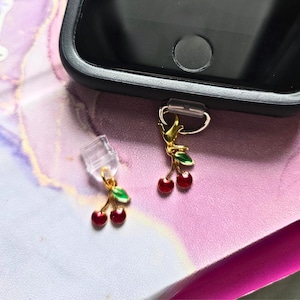 May include: Two cherry phone charms with red enamel cherries and green leaves, attached to gold-colored metal clasps. One charm is connected to a black phone case, the other is detached. The background is a pink and white marbled pattern.