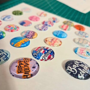 May include: A collection of oval-shaped, clear-domed magnets with various designs and text. Designs include patriotic themes, holiday greetings like "Happy Halloween," and seasonal messages such as "Summer" and "Back to School."