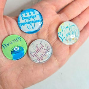 May include: Four round, clear glass magnets with various designs. One magnet says "Dentist Appointment" in blue, another says "Therapy" in green, a third says "Bridal Shower" in pink, and the last says "Baby Shower" in blue and yellow.
