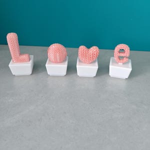 Peut inclure: Quatre lettres roses tricotées épellent le mot "LOVE", présentées sur des bases carrées blanches individuelles. Les lettres ont un aspect texturé, et le fond est de couleur turquoise. L'arrangement est sur une surface grise.