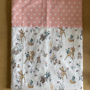 May include: A folded, rectangular fabric with a two-tone design. The top section features a light pink background with white stars. The bottom section has a white background with a pattern of deer, squirrels, and other forest creatures.