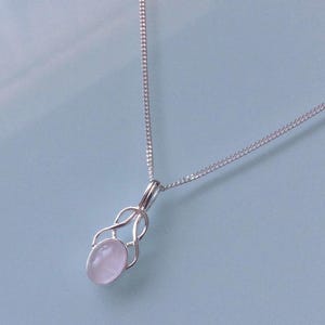 May include: A silver necklace with a fine chain and a pendant. The pendant holds a pale pink oval gemstone, framed by an openwork design. The necklace has a simple, elegant style, suitable for everyday wear or special occasions.