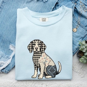 May include: A light blue crew neck t-shirt featuring a graphic of a sitting dog. The dog's body is divided into sections with different patterns, including black and white buffalo check, a floral print, polka dots, and a dark blue damask design.
