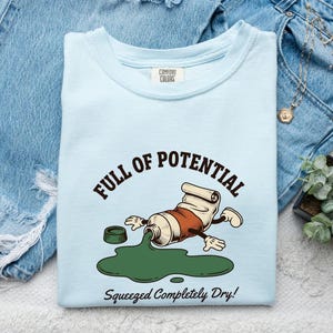 Op de afbeelding: Een lichtblauw t-shirt met een afbeelding van een cartoon verftube met armen en benen, waar groene verf uit lekt. De tekst luidt FULL OF POTENTIAL boven de tube en Squeezed Completely Dry! eronder. Het shirt heeft een Comfort Colors merklabel.