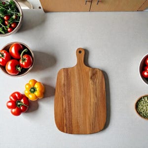以下が含まれることがあります： ハンドル付きの木製まな板が新鮮な食材に囲まれています。赤いトマトとパプリカ、そしてグリーンのボウルが明るい色のカウンタートップに配置されています。まな板は明るい茶色の木材で作られています。
