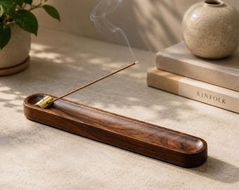 May include: A long, rectangular dark wood incense holder with a smooth, polished finish. A small, cylindrical brass insert is positioned at one end, holding a single thin incense stick that emits a delicate, swirling trail of white smoke.