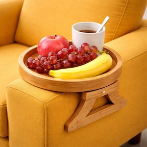 Puede incluir: Una bandeja de madera para el brazo del sofá contiene una manzana roja, un racimo de uvas rojas y un plátano amarillo. Una taza blanca con una cuchara y un líquido oscuro se encuentra en la bandeja. La bandeja está sujeta al brazo de un sofá amarillo.