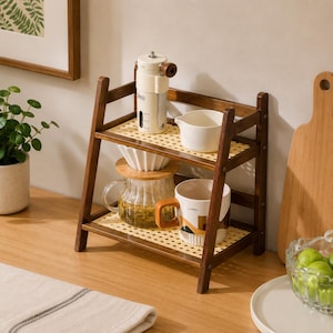 May include: A two-tier wooden storage shelf with a light-coloured woven cane base on each level. The top shelf holds a white manual coffee grinder and a small white ceramic pitcher. The bottom shelf holds a glass coffee carafe and a ceramic mug.